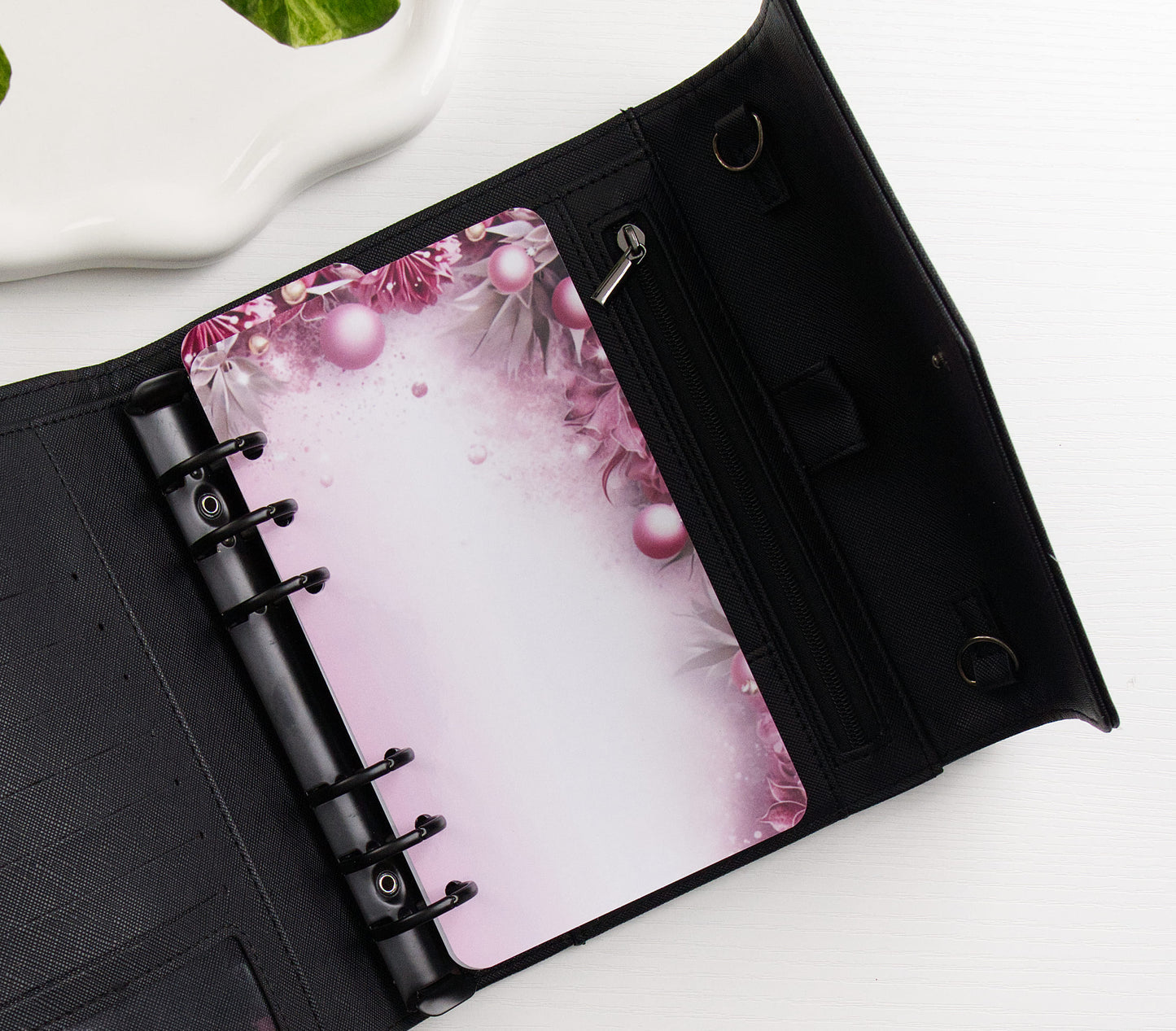 A black ring binder shown open on a white surface, featuring a pink floral and pearl-patterned dashboard page.