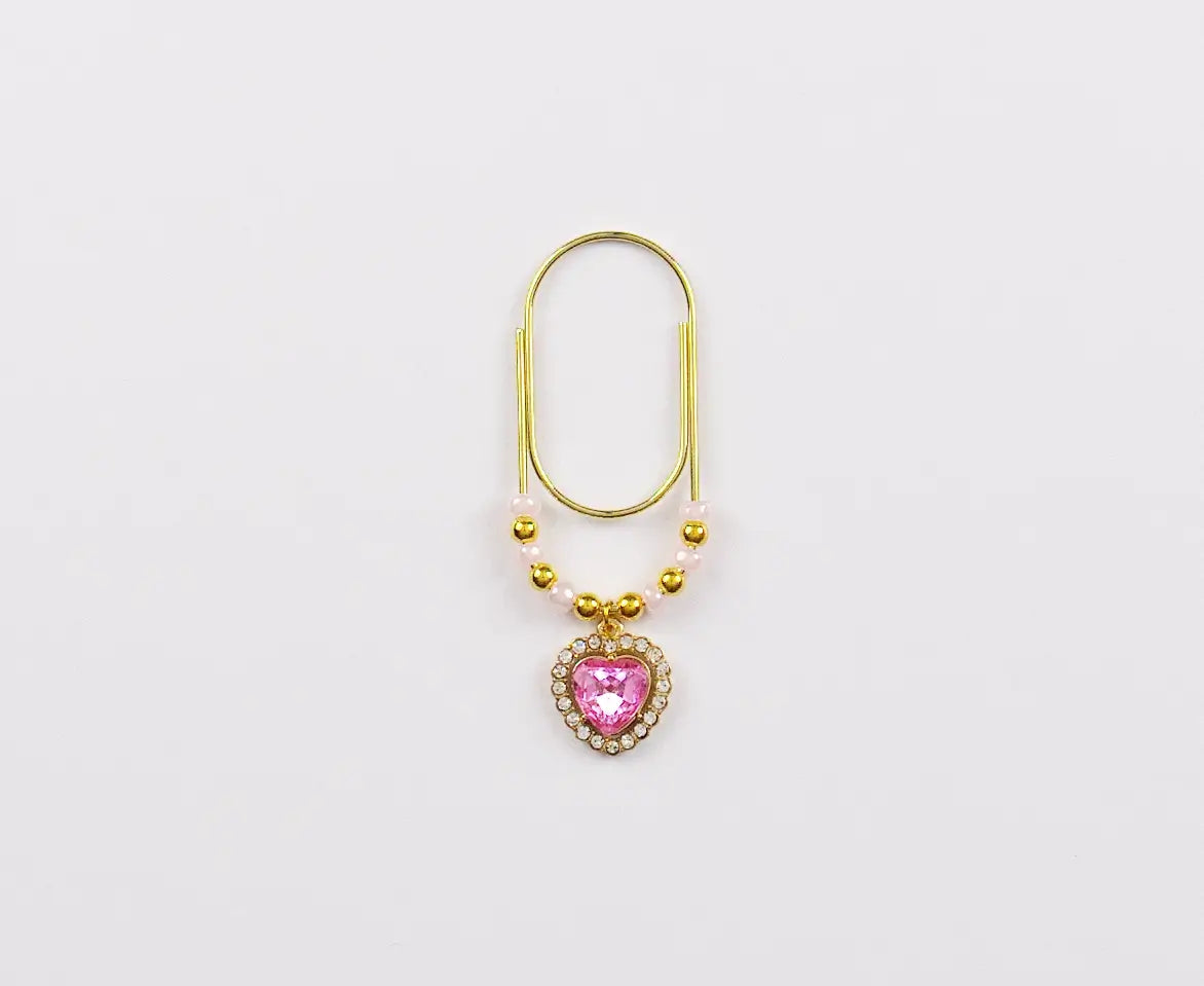 Gold paper clip charm with a pink heart-shaped gem on a white background