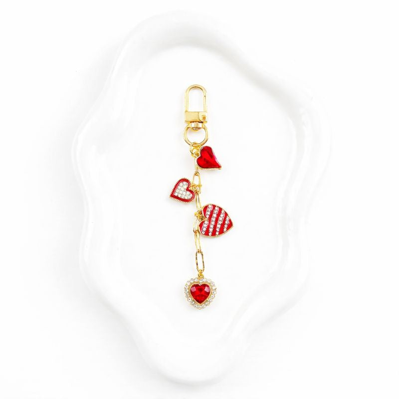 Gold binder charm with cascading red heart pendants featuring enamel, rhinestone, and striped designs on paperclip chain with lobster clasp, displayed on white wavy trinket dish against sage green background