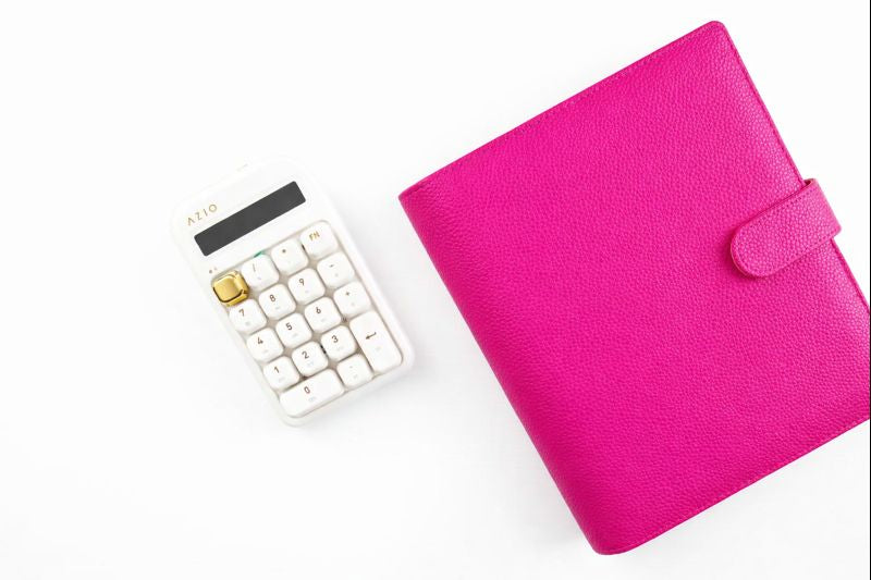 Hot pink pebbled leather A6 6-ring binder with snap tab closure displayed closed on white background beside white  calculator with gold accent, showcasing textured finish