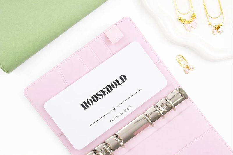 Minimalist white Household cash envelope insert with elegant black typography tucked into card slot of blush pink A6 binder with silver rings, styled with sage green binder and gold bow charms on trinket dish