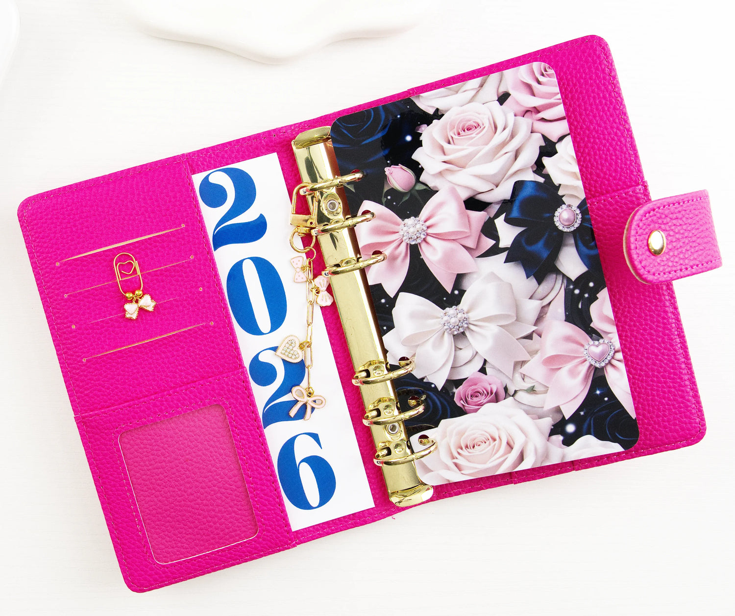 Hot pink pebbled leather A6 binder opened flat showing gold rings, card slots, ID window, 2026 dashboard insert, and floral divider with pink roses and satin bows, decorated with gold bow and heart binder charms