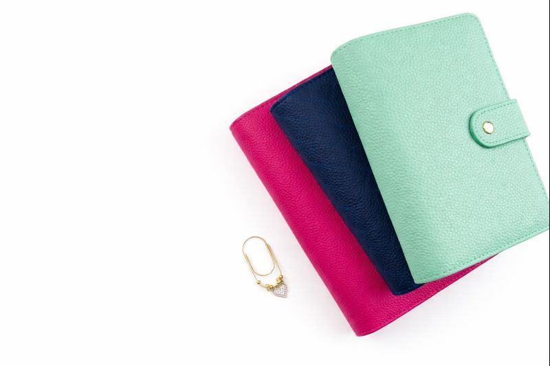 Three pebbled leather A6 binders stacked in mint green, navy, and hot pink with gold snap closures, displayed on white background with gold heart binder charm