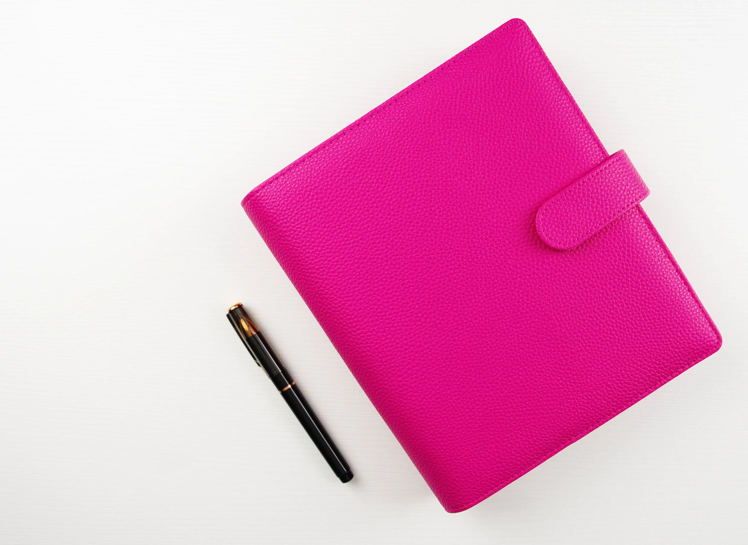 Hot pink pebbled leather A5 6-ring binder with snap tab closure displayed closed on white background beside black and gold fountain pen, showcasing textured finish and premium construction