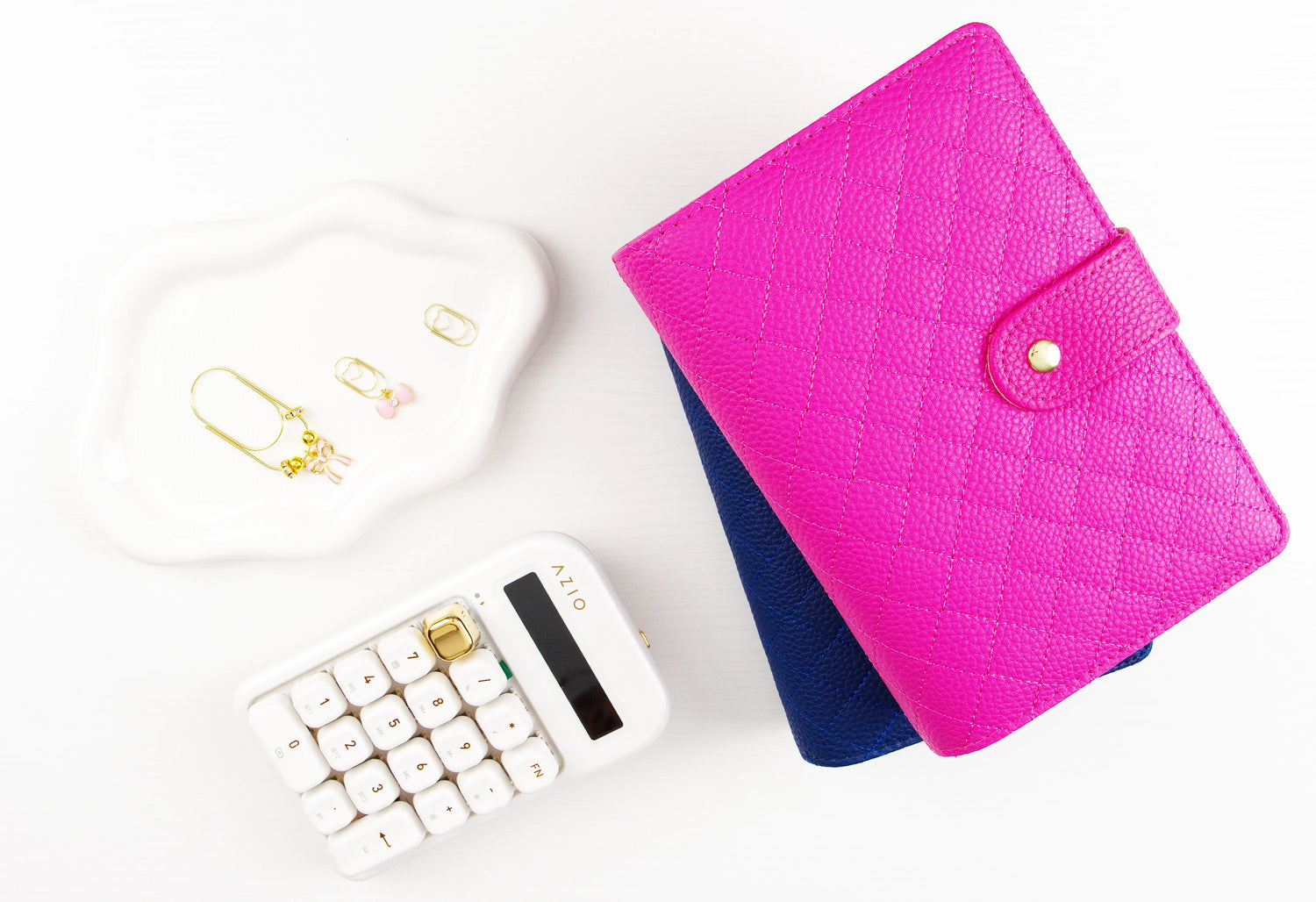 Flat lay of hot pink quilted pebbled leather A6 binder with gold snap closure stacked on navy binder, styled with white calculator, gold binder charms, and trinket dish on white background