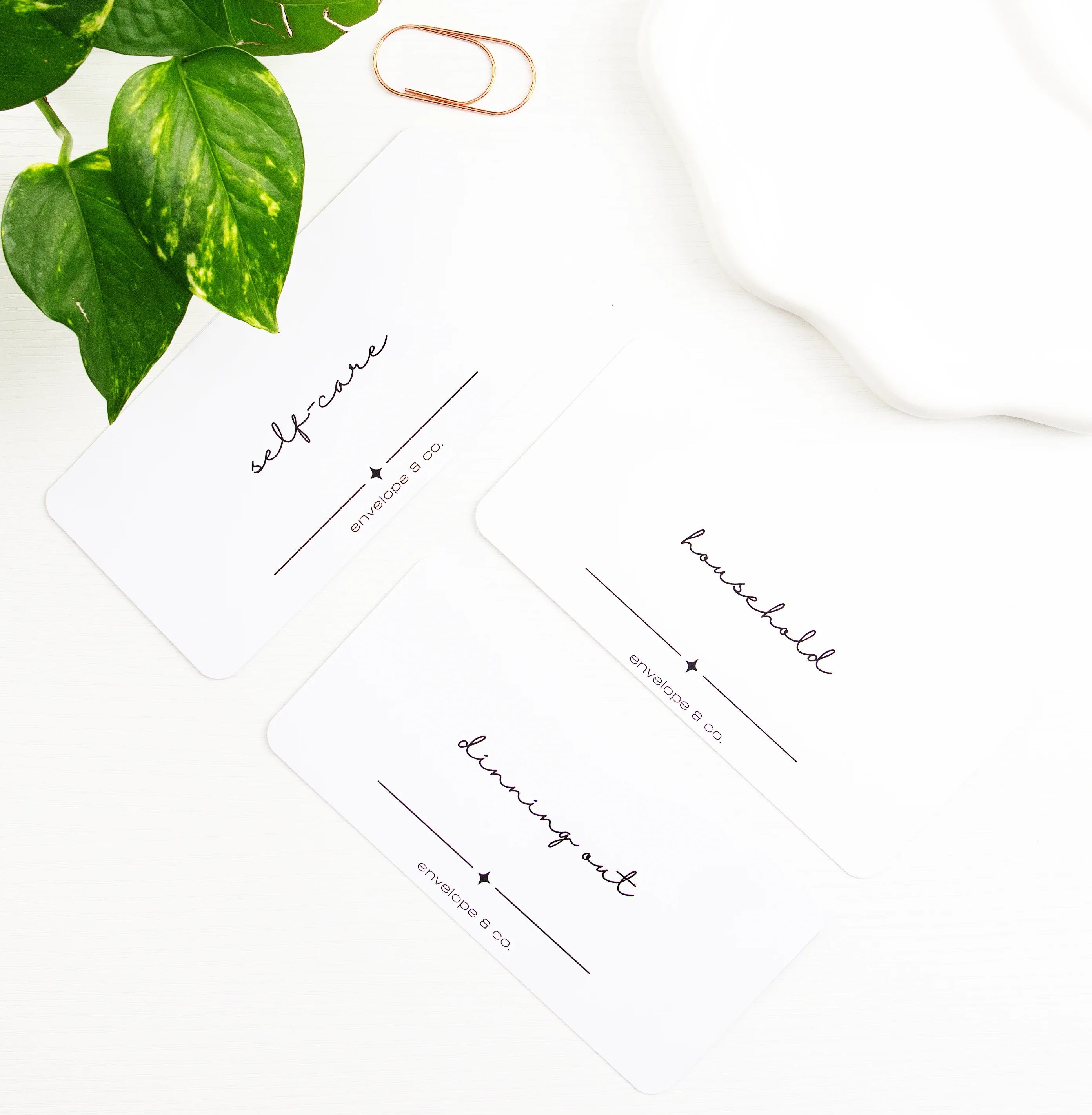 Three minimalist white cash envelope inserts with elegant script labels reading Self-Care, Household, and Dining Out, styled flat lay with pothos plant, rose gold paper clip, and white trinket dish