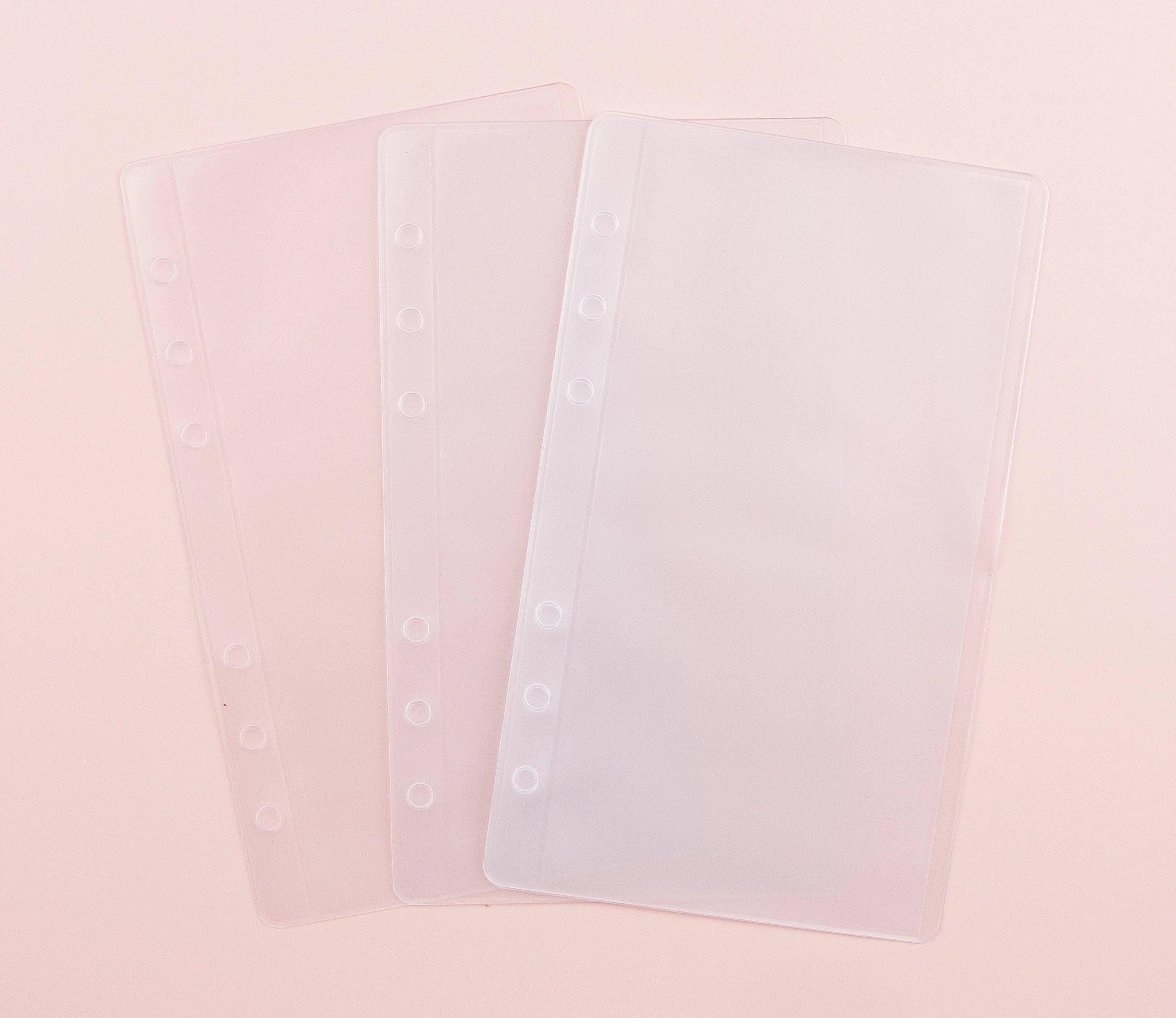Two clear frosted plastic cash envelopes with 6-hole punch for A6 ring binders, featuring top-loading pocket design, displayed overlapping on blush pink background