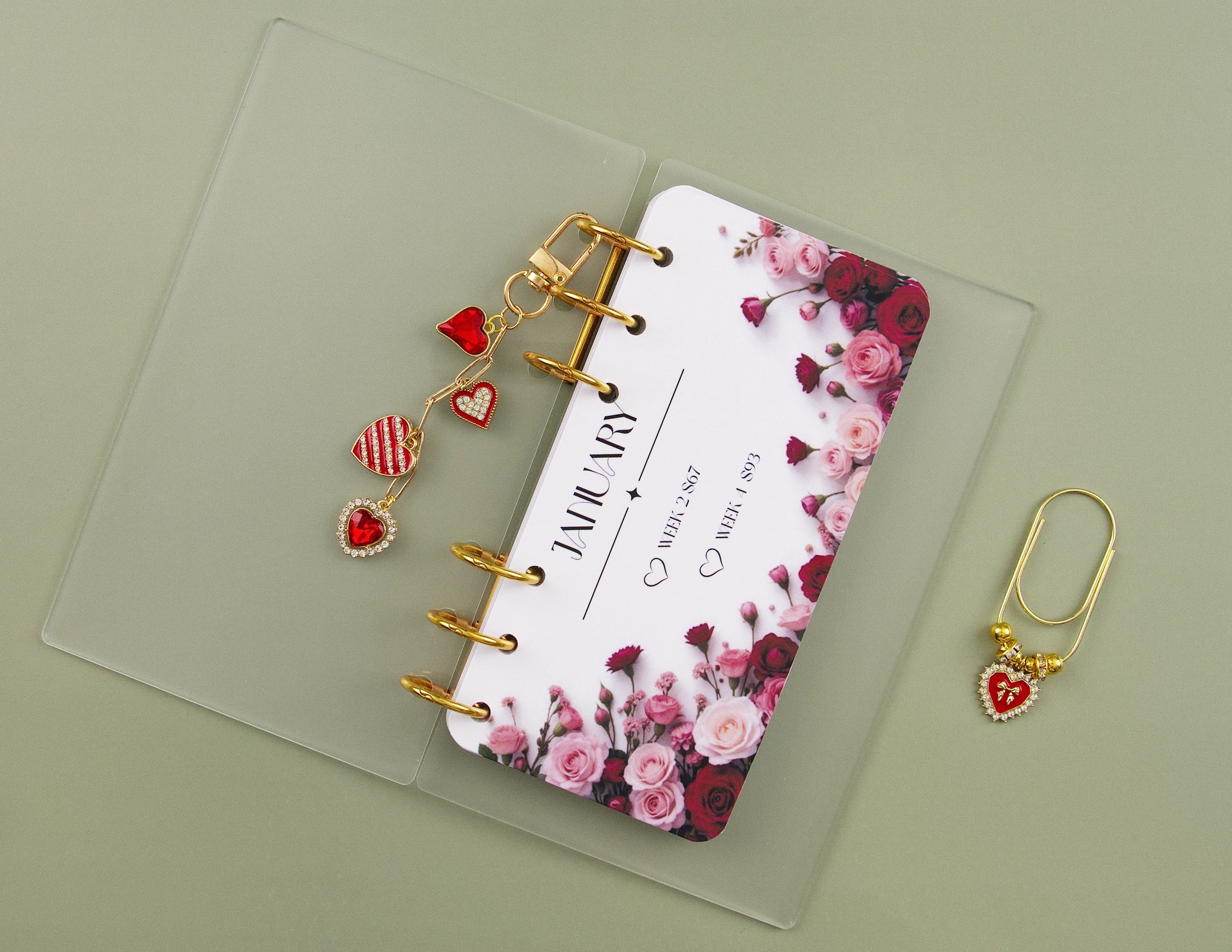 Clear acrylic A6 binder with gold rings showing January savings tracker with pink and red roses, accompanied by two red heart binder charms