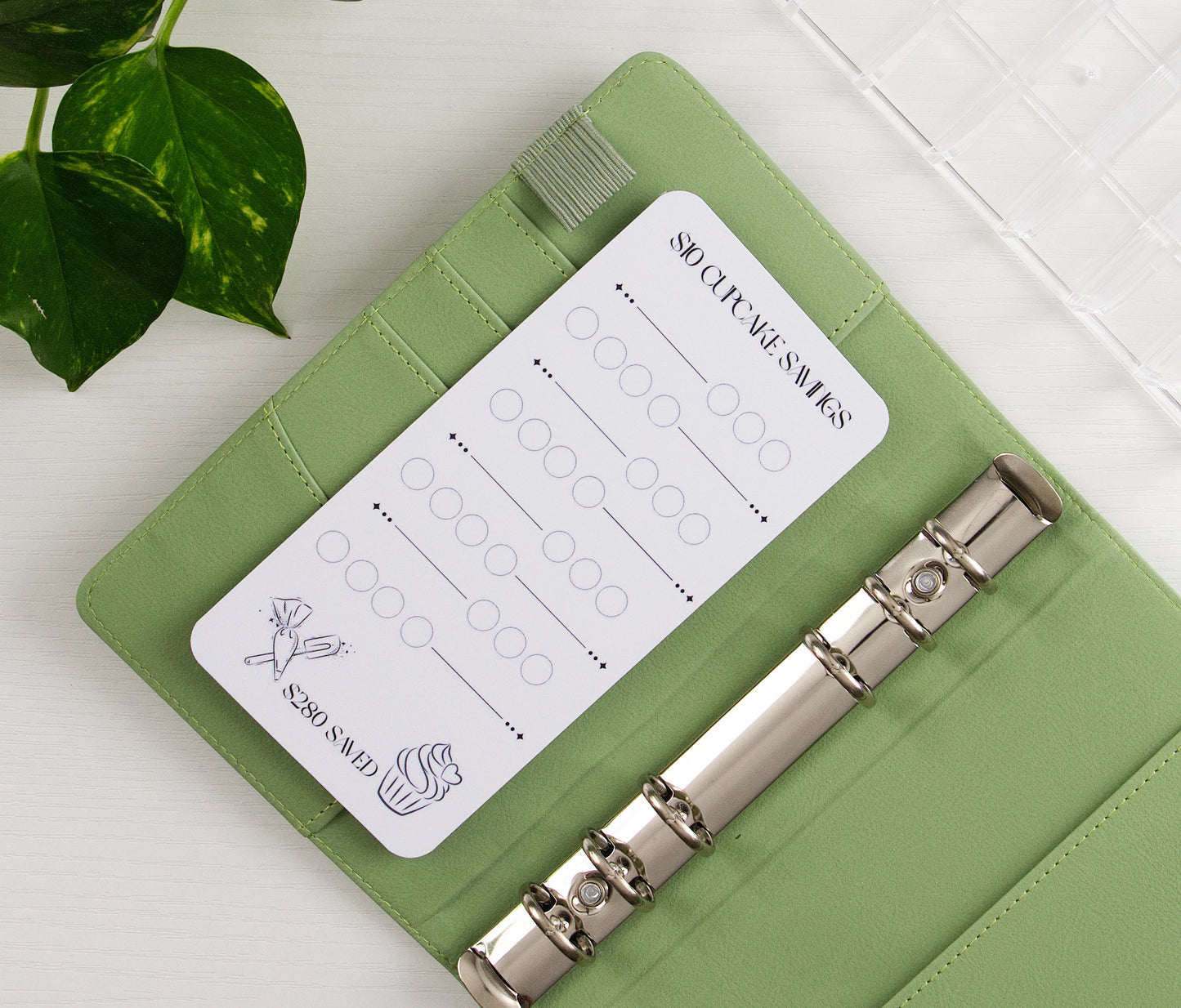 $10 Cupcake Savings challenge with circles to track $10 deposits toward $280 goal, featuring cupcake and party icons in a sage green A6 binder with silver rings beside a plant