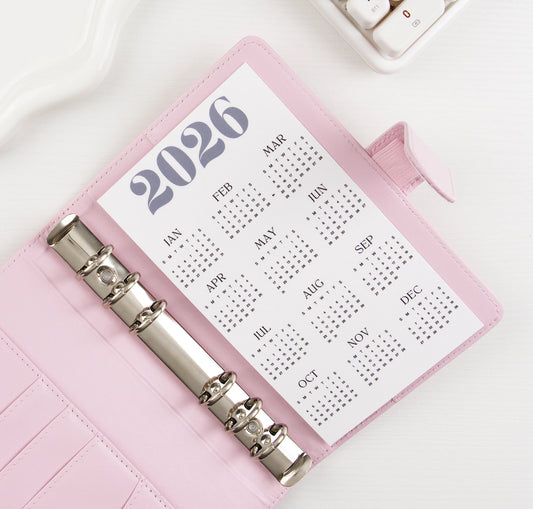 2026 yearly calendar dashboard insert with navy blue header and 12-month grid layout displayed in open blush pink A6 binder with silver rings, styled beside white calculator