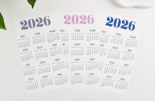 Three 2026 yearly calendar dashboard inserts displayed overlapping, featuring minimalist design with 12-month grid layout in navy, mauve, and dusty blue color options, styled on white background with plant accent