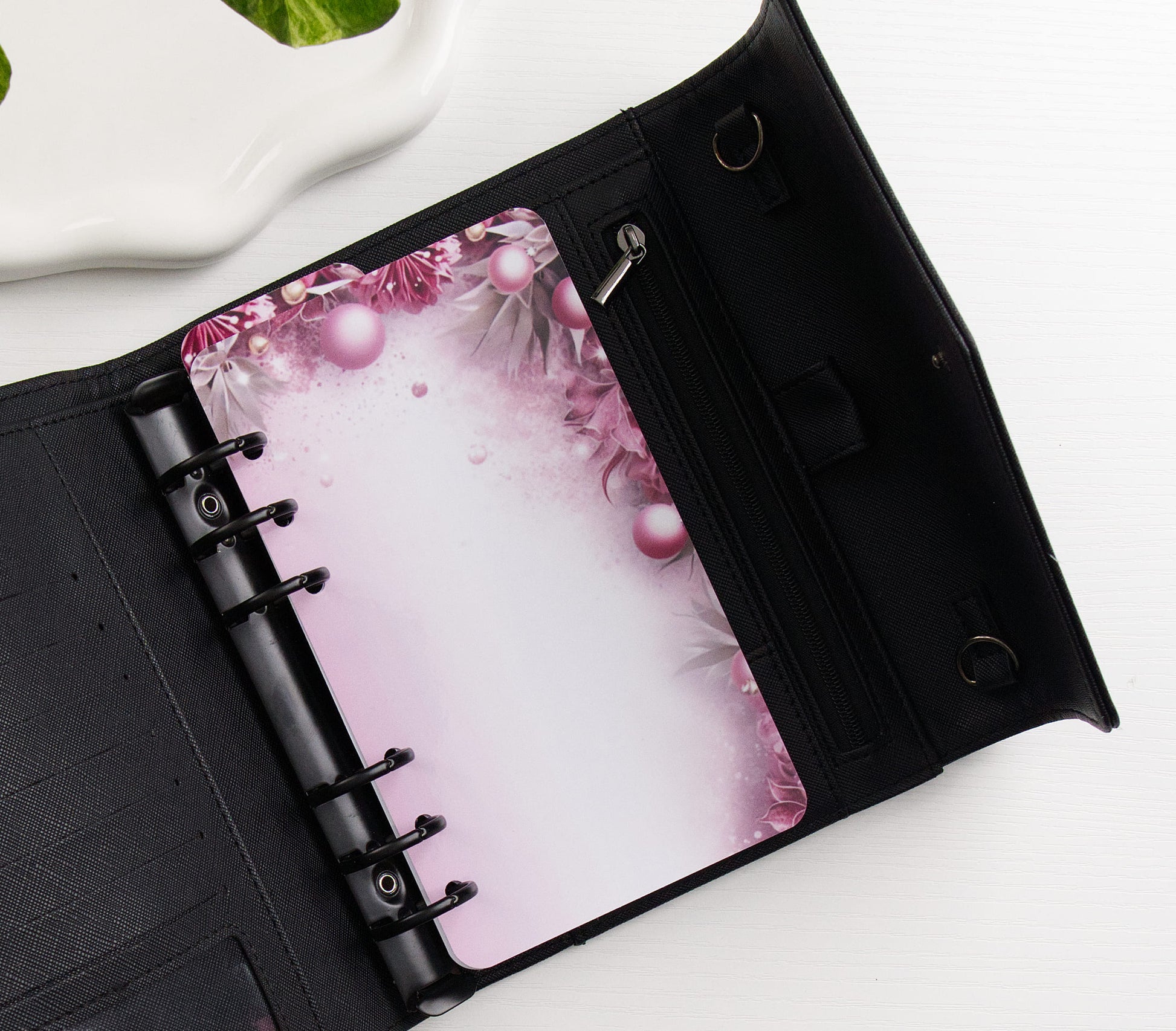 A black ring binder shown open on a white surface, featuring a pink floral and pearl-patterned dashboard page.