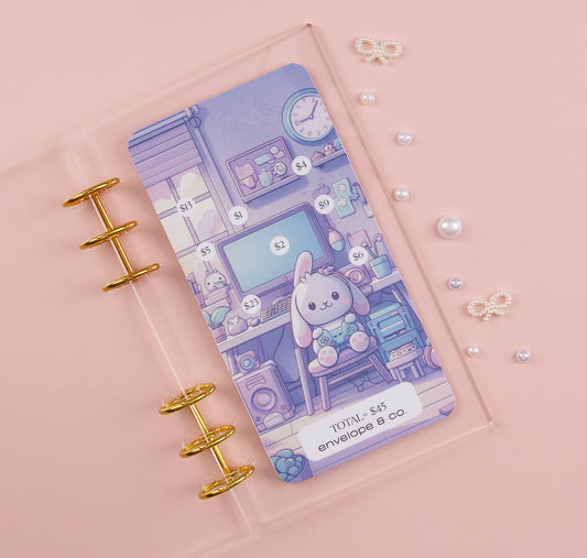 Kawaii-style savings challenge insert featuring pastel purple illustration of cute bunny in cozy gaming room, with dollar amounts from $1-$23 scattered throughout scene to mark off, $45 total goal, shown in clear A6 binder with gold rings on pink background