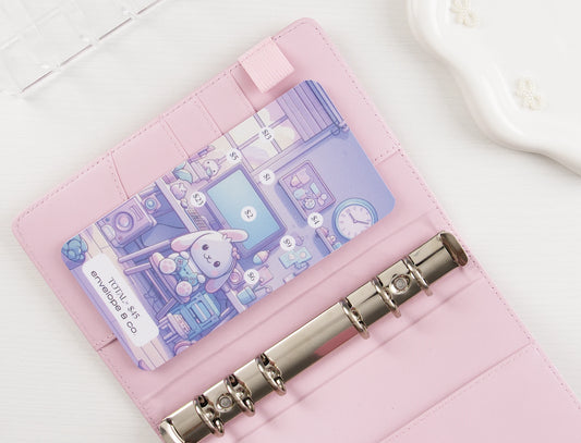Kawaii savings challenge insert with pastel purple bunny gaming room illustration, dollar amounts $1-$13 scattered throughout scene toward $45 goal, tucked into card slot of blush pink A6 binder with silver rings