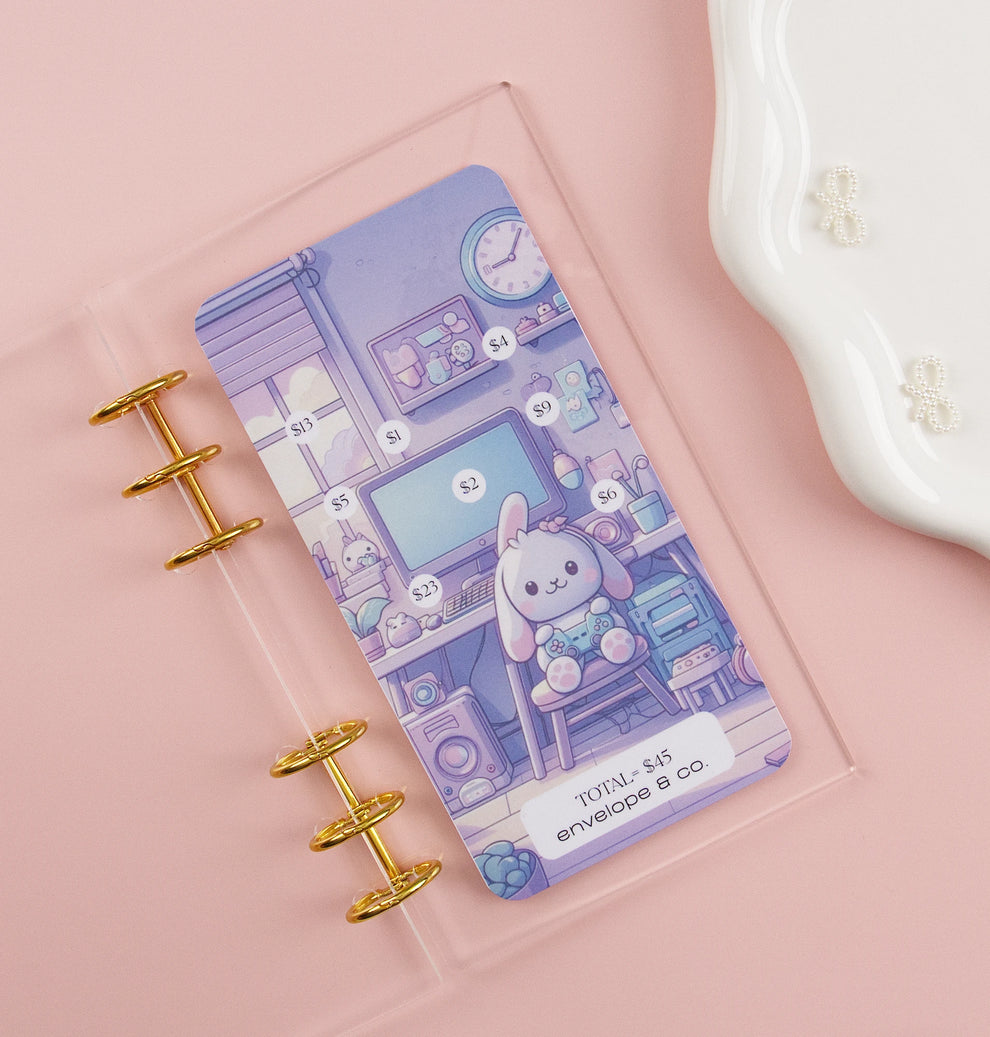 Kawaii savings challenge insert featuring pastel purple bunny gaming room illustration with dollar amounts $1-$23 to mark off toward $45 goal, displayed in clear A6 binder with gold rings on pink background