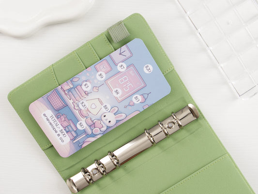Kawaii savings challenge insert with pastel purple retro gaming illustration featuring handheld consoles and controllers, dollar amounts $1-$20 to mark off toward $100 goal, tucked into sage green A6 binder with silver rings