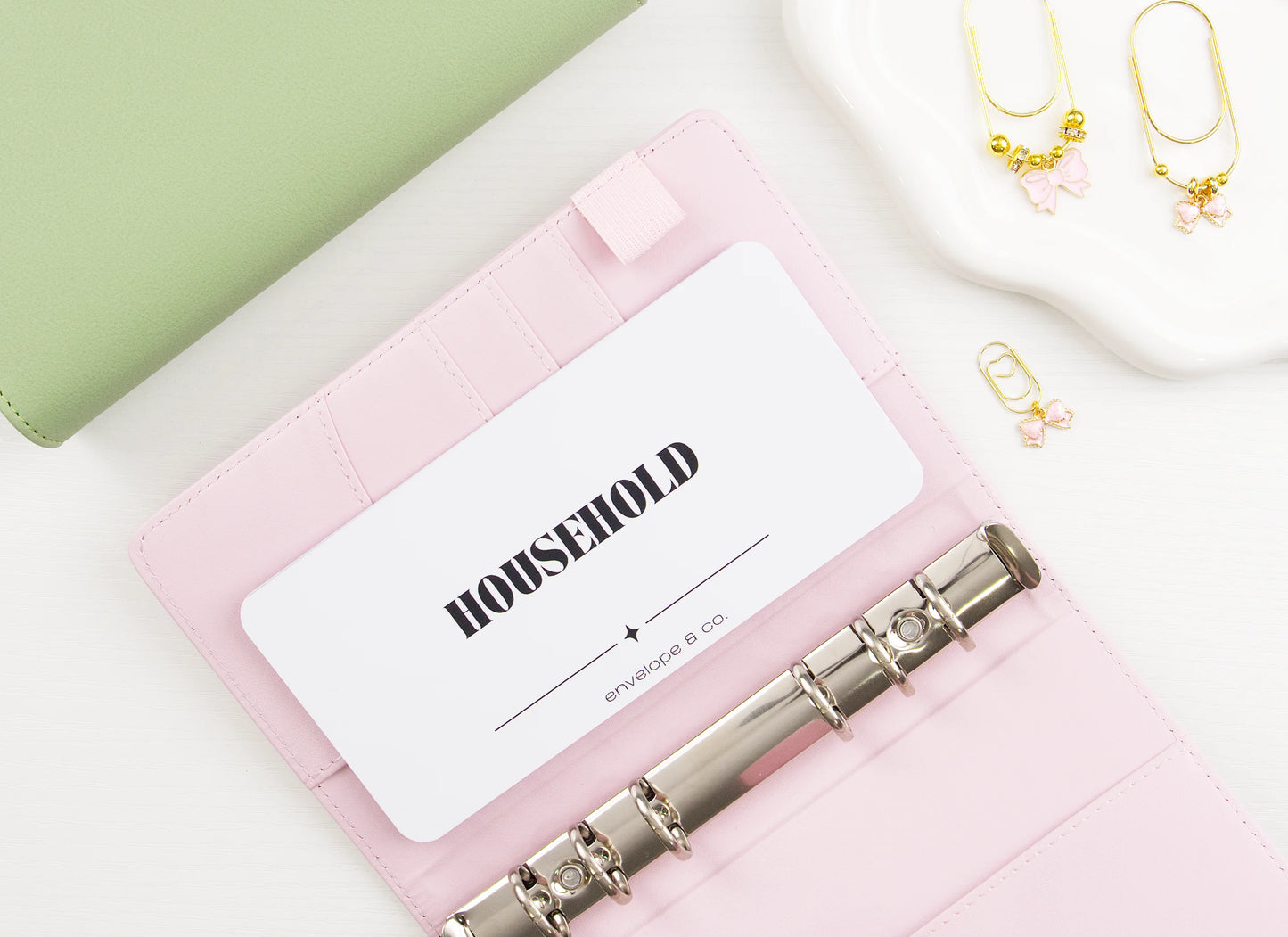 Minimalist white Household cash envelope insert with elegant black typography tucked into card slot of blush pink A6 binder with silver rings, styled with sage green binder and gold bow charms on trinket dish