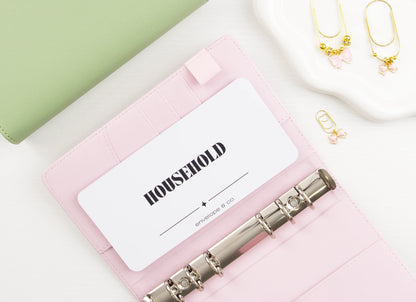 Minimalist white Household cash envelope insert with elegant black typography tucked into card slot of blush pink A6 binder with silver rings, styled with sage green binder and gold bow charms on trinket dish