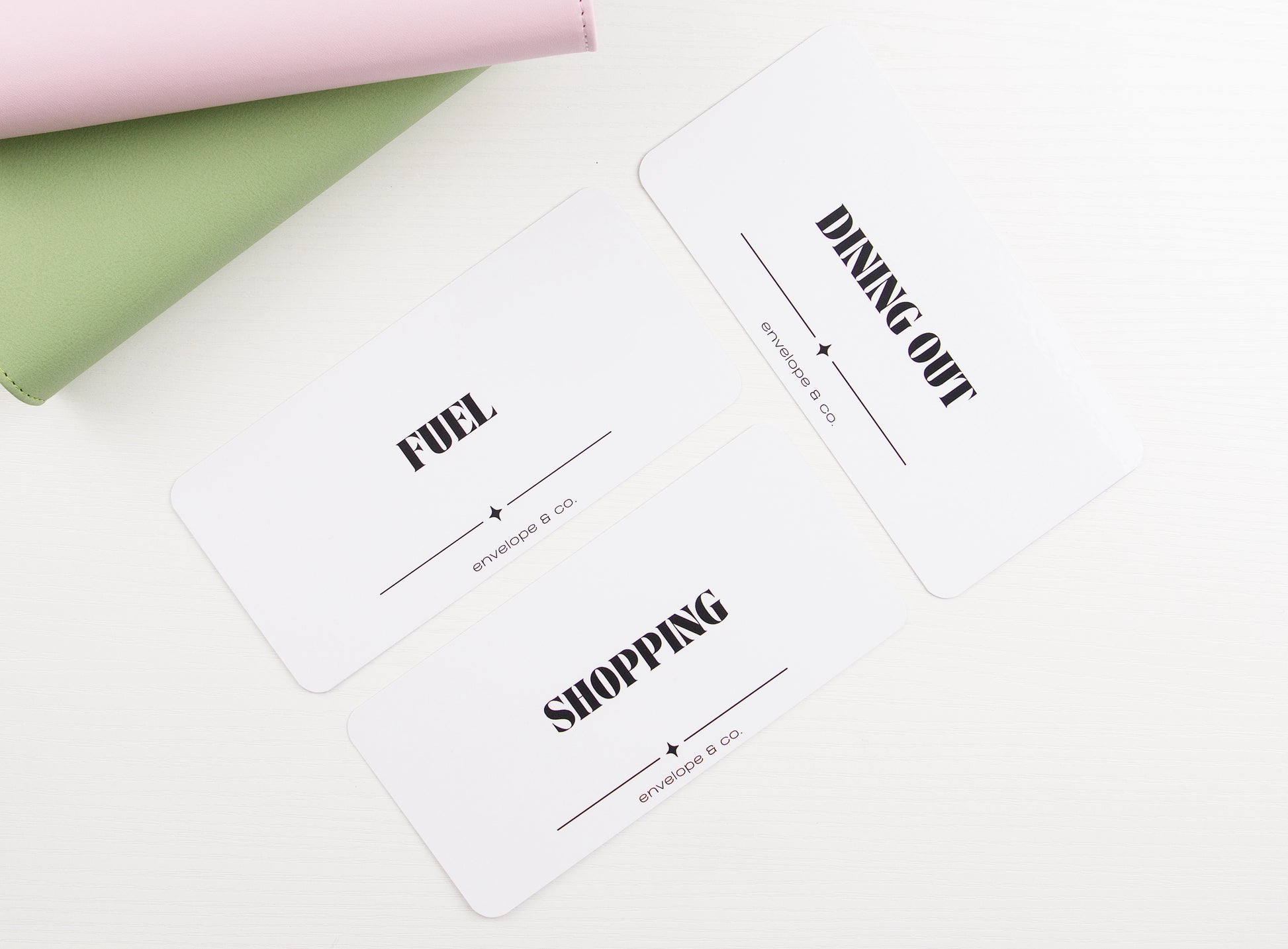 Three minimalist white cash envelope inserts with bold black typography reading Fuel, Dining Out, and Shopping, styled with blush pink and sage green binders on white background