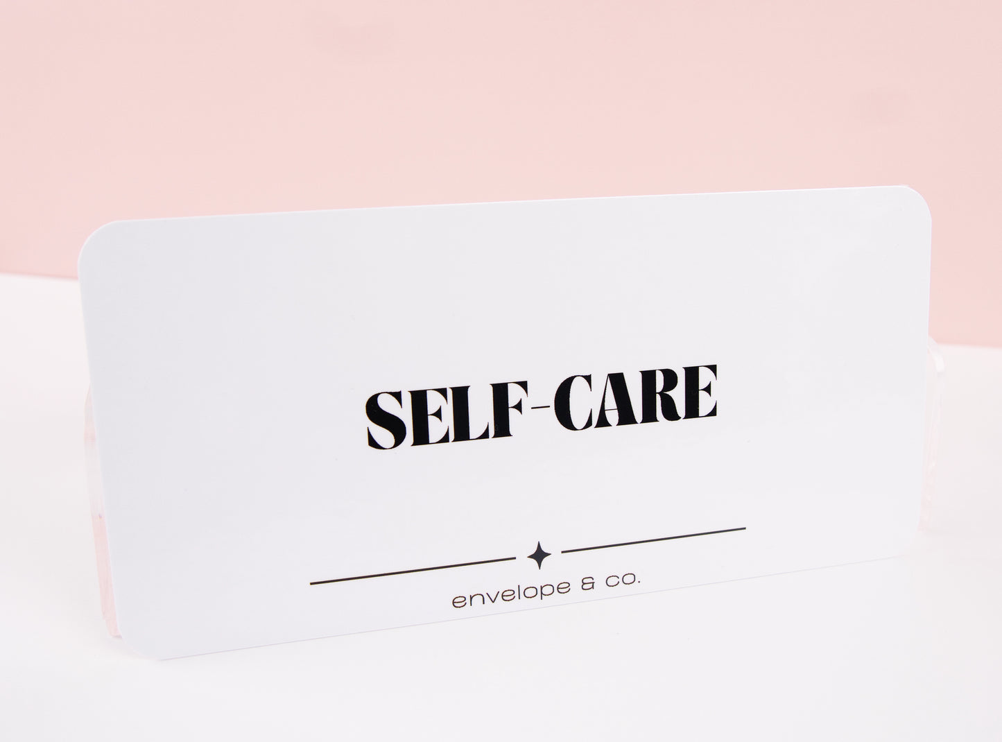 Minimalist white Self-Care cash envelope insert with bold black typography and star accent line, displayed standing against soft pink background
