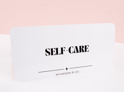 Minimalist white Self-Care cash envelope insert with bold black typography and star accent line, displayed standing against soft pink background
