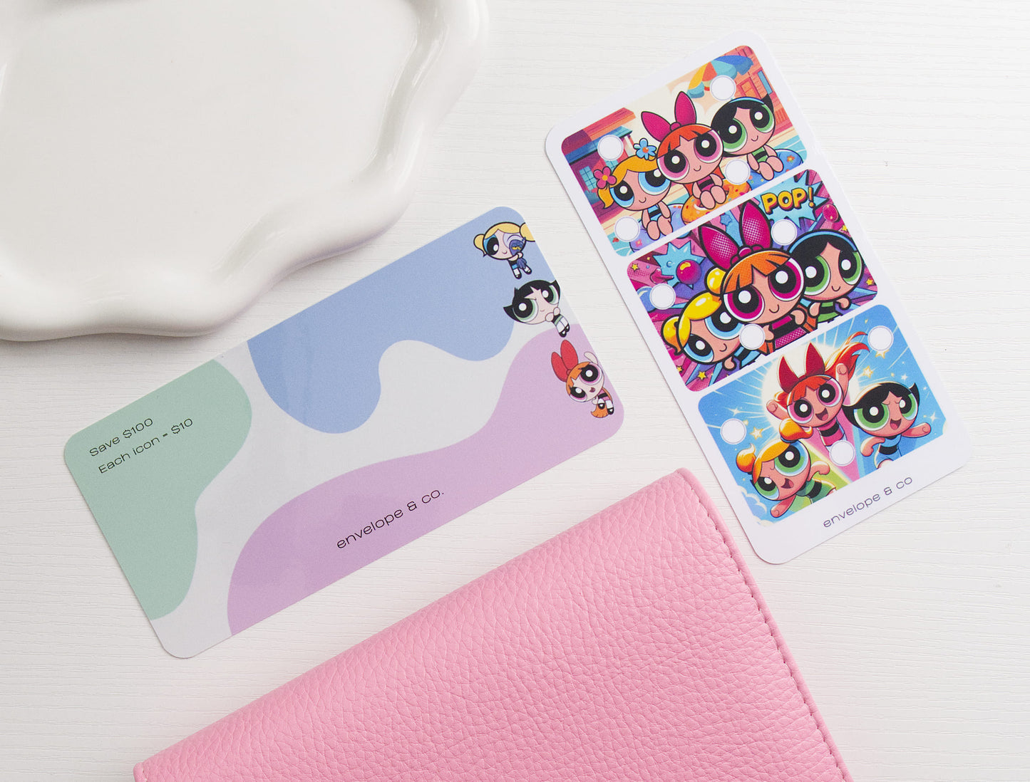 A set of colorful dashboard and savings challenge displayed on a white textured surface next to a pink textured binder and a white ceramic dish.