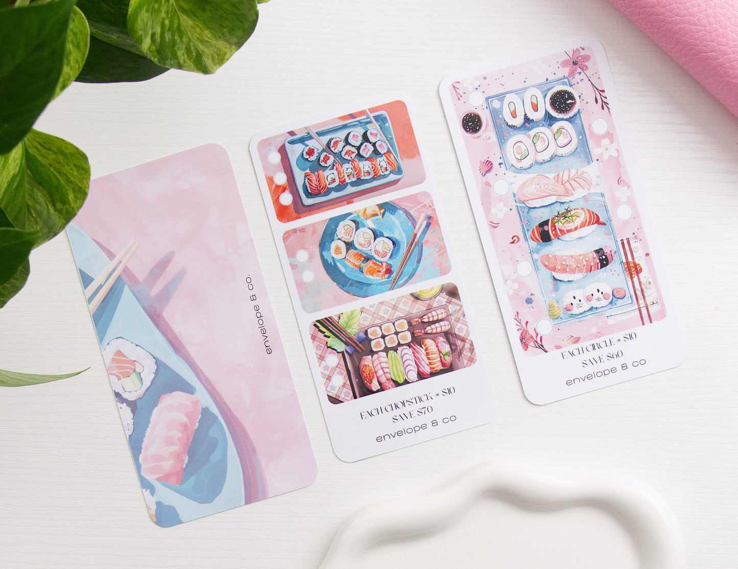 Two sushi themed savings challenge and one dashboard arranged on a white surface, alongside green leaves and a pink binder.