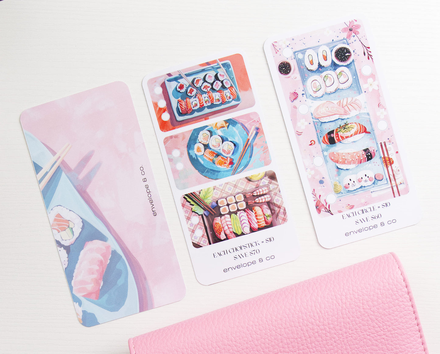Two sushi themed savings challenge and one dashboard arranged on a white surface, alongside a pink binder on a white background.