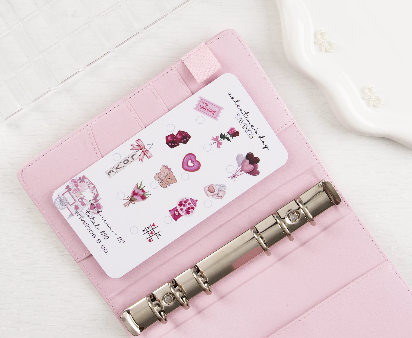 Valentine's Day Savings challenge insert with pink romantic icons including hearts, roses, teddy bear, chocolates, and balloons to mark off toward $110 goal, tucked into blush pink A6 binder with silver rings