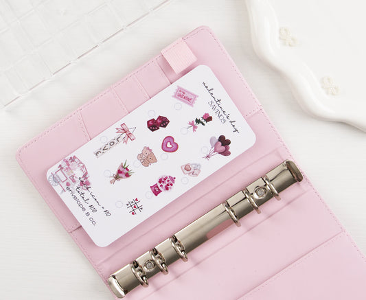 Valentine's Day Savings challenge insert with pink romantic icons including hearts, roses, teddy bear, chocolates, and balloons to mark off toward $110 goal, tucked into blush pink A6 binder with silver rings