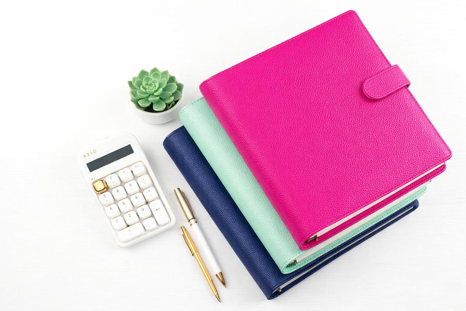 Stack of three pebbled leather A5 binders in hot pink, mint green, and navy blue with snap closures, styled on white background with white calculator, gold pens, and small succulent plant