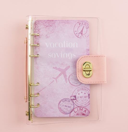 Pink clear acrylic A6 binder with gold rings showing vacation savings dashboard with passport stamps and travel imagery on pink background