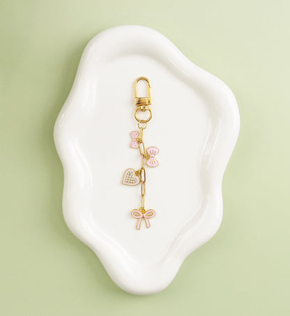 Gold heart-themed charm with pink enamel hearts, displayed on a white dish against a green background.