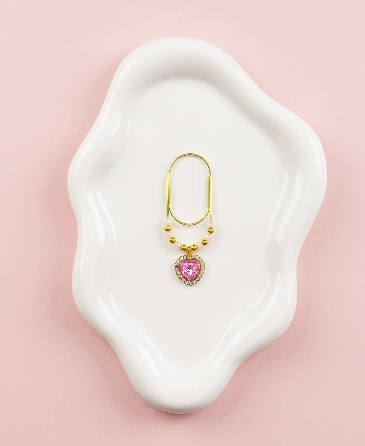 Gold paper clip charm with a pink gemstone on a white dish against a pink background