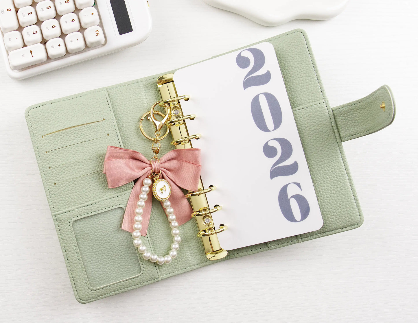 Green binder with '2026' on a white surface, featuring decorative elements like a pink bow and pearl charm.