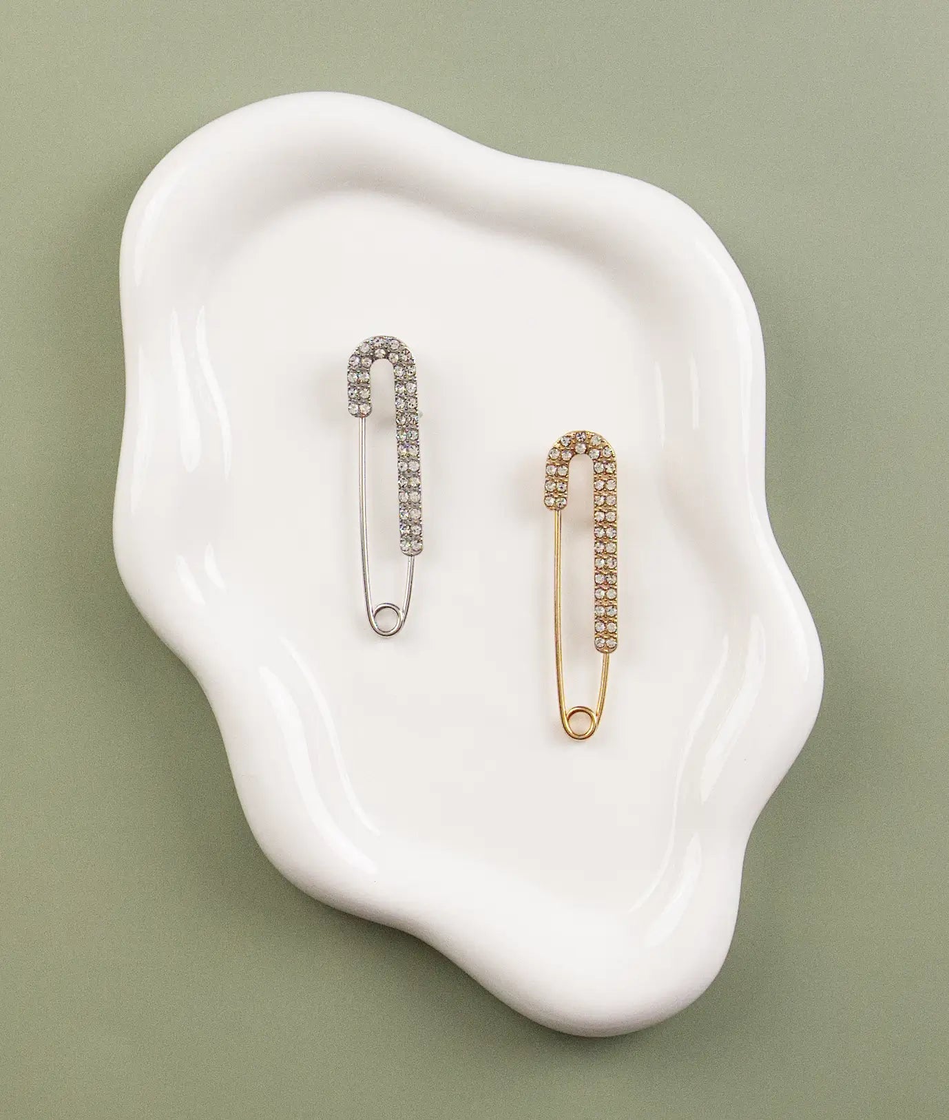Two decorative safety pin charms with gemstones on a white dish against a green background