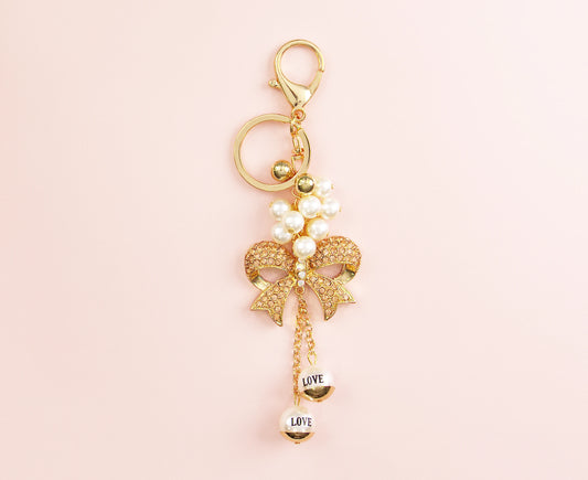 Gold bow charm with pearls and dangling “LOVE” beads, clipped to a key ring on a pink background.