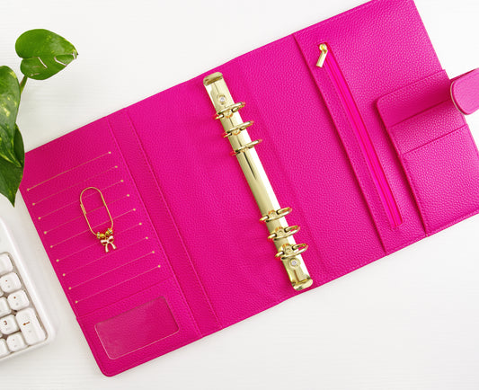 Pink binder with gold binding and a gold paper clip with pink bow charm on a white surface