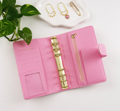 Pink binder with gold accents on a white surface, with earrings in the background.