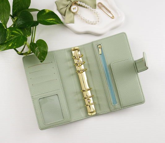 Sage green binder with gold accents on a white surface with a plant in the corner.