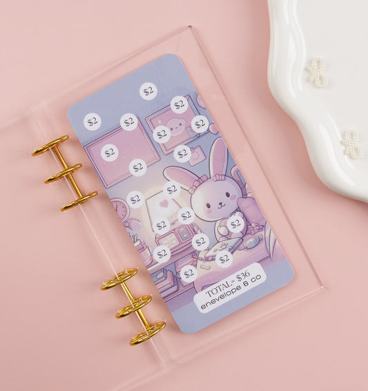 Kawaii bunny savings tracker with $2 increments totaling $36 displayed in clear acrylic binder with gold rings on pink background