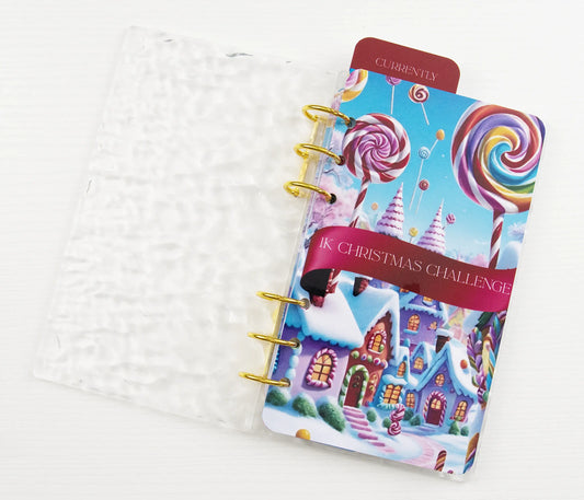 Clear acrylic binder with gold rings showing 1K Christmas Challenge tracker featuring colorful candy canes and gingerbread houses.