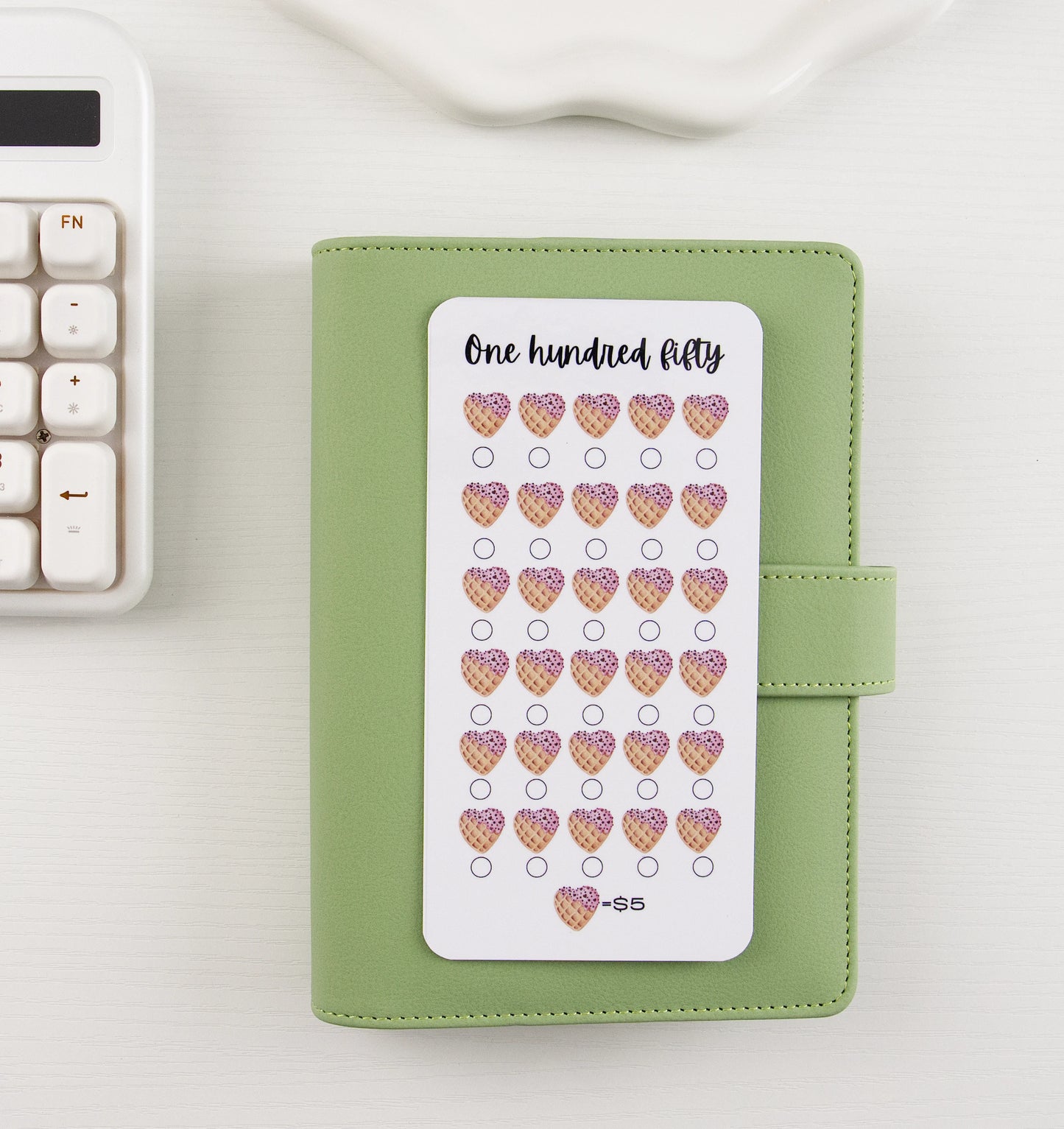 One Hundred Fifty dollar savings tracker with 30 ice cream waffle cone hearts at $5 each displayed on top of a sage green binder on white background
