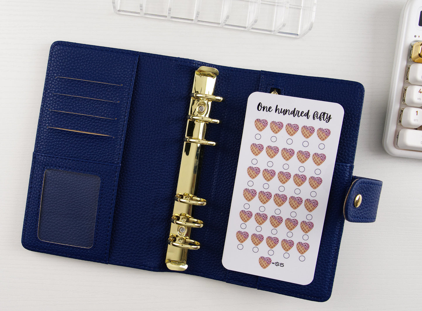 Open navy blue vegan leather A6 binder with gold rings showing One Hundred Fifty dollar savings tracker with ice cream cone hearts on white background